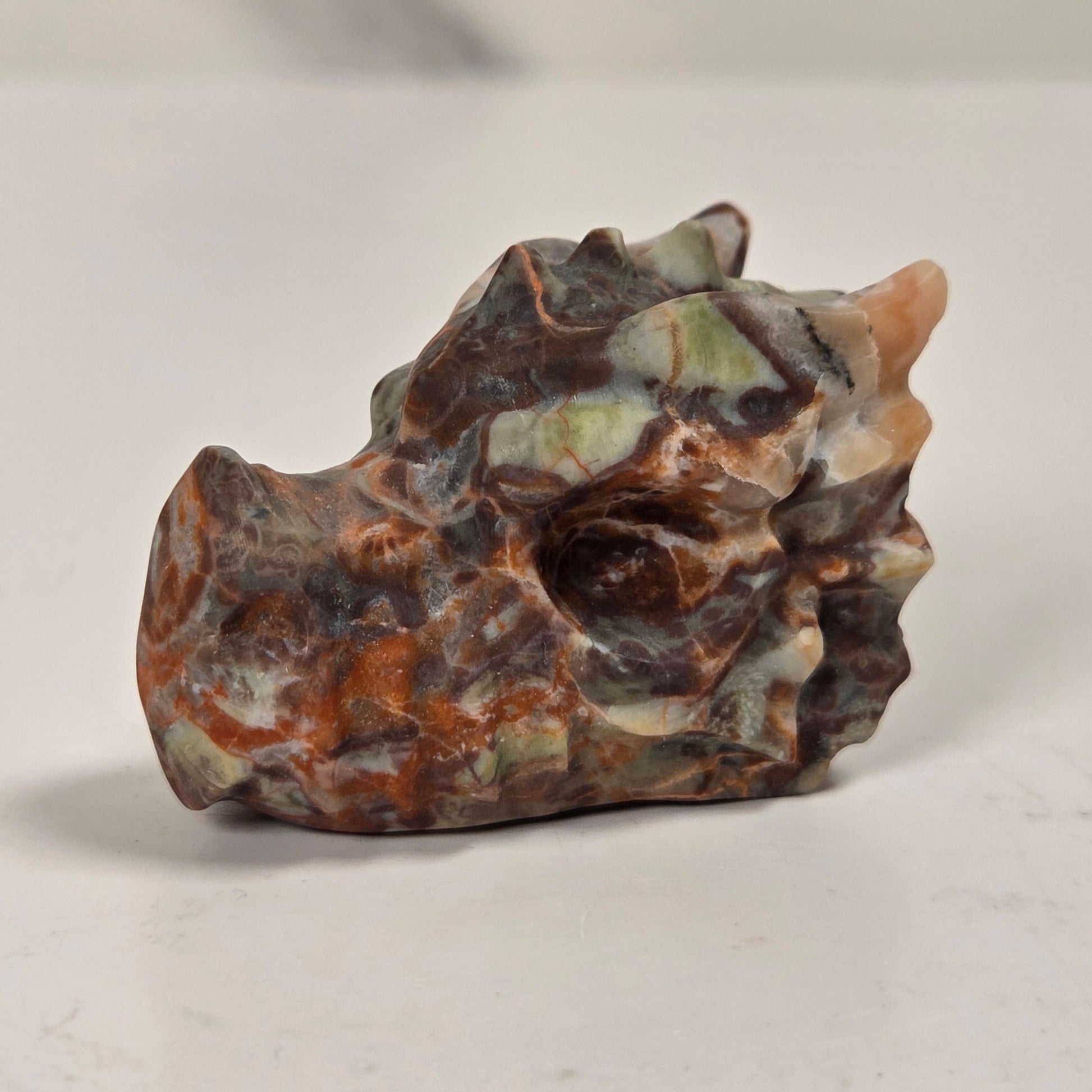 A small carved stone sculpture resembling a dragon head, featuring natural marbled textures in brown, orange, green, and gray, displayed on a light gray surface.