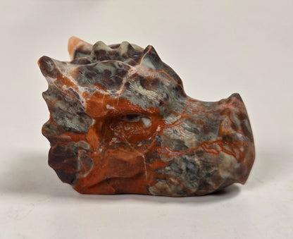 A small carved stone figurine resembling a stylized animal head or creature, made from polished natural stone with mottled orange, brown, gray, and cream colors, standing on a light gray surface.