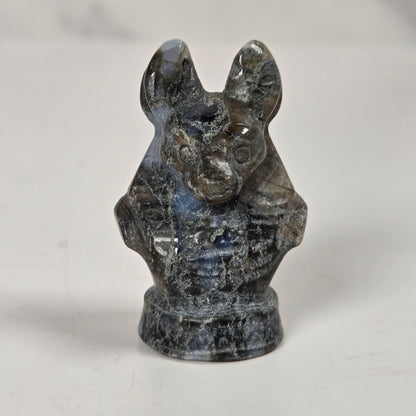 A small carved stone figurine of a bat, likely made of labradorite, featuring a mottled dark gray and blue-gray coloration with natural crystalline inclusions, standing on a small circular base against a light gray background.