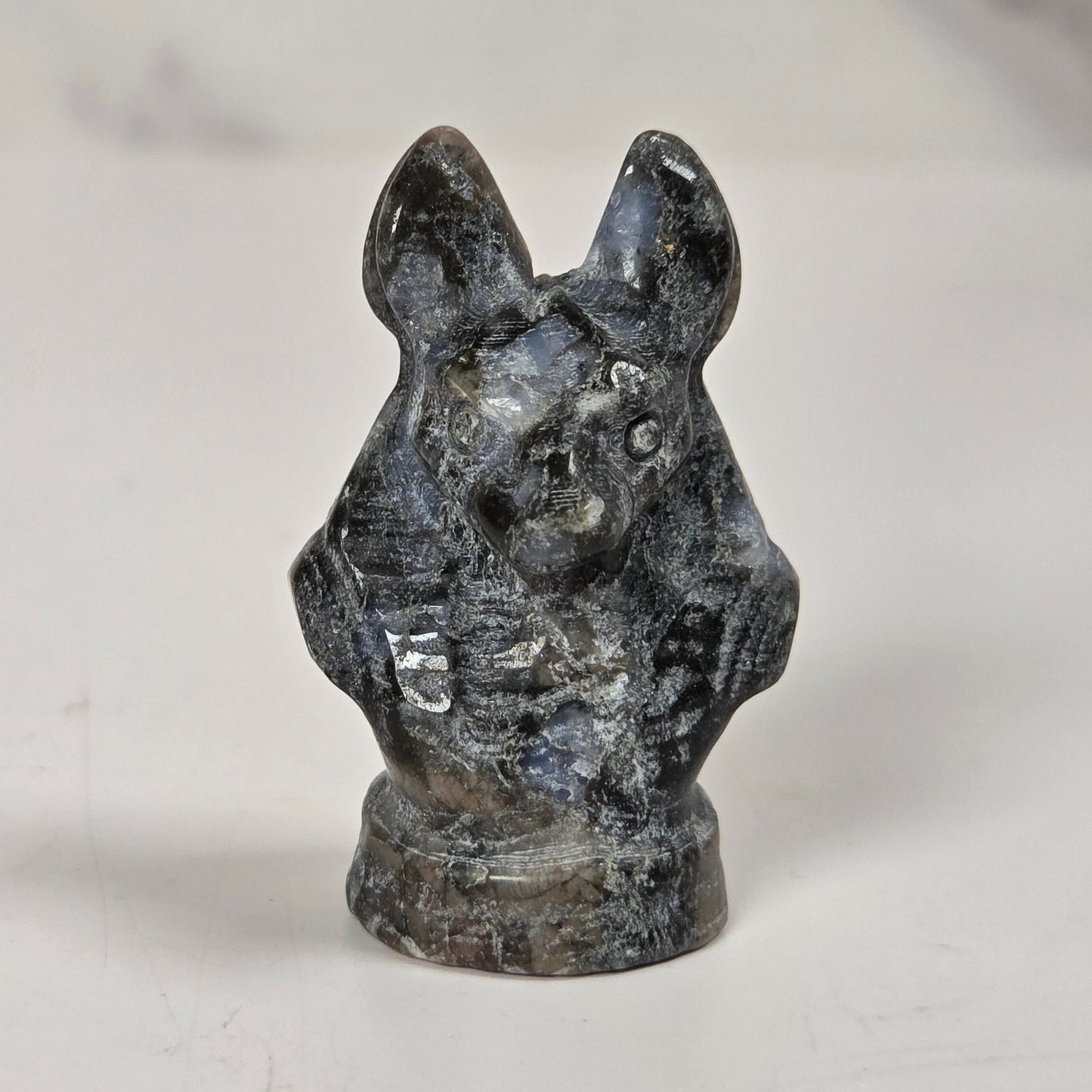 A small carved figurine of a rabbit or hare head and upper body, made from a dark polished stone with gray, black, and blue-gray marbling, standing on a white surface.