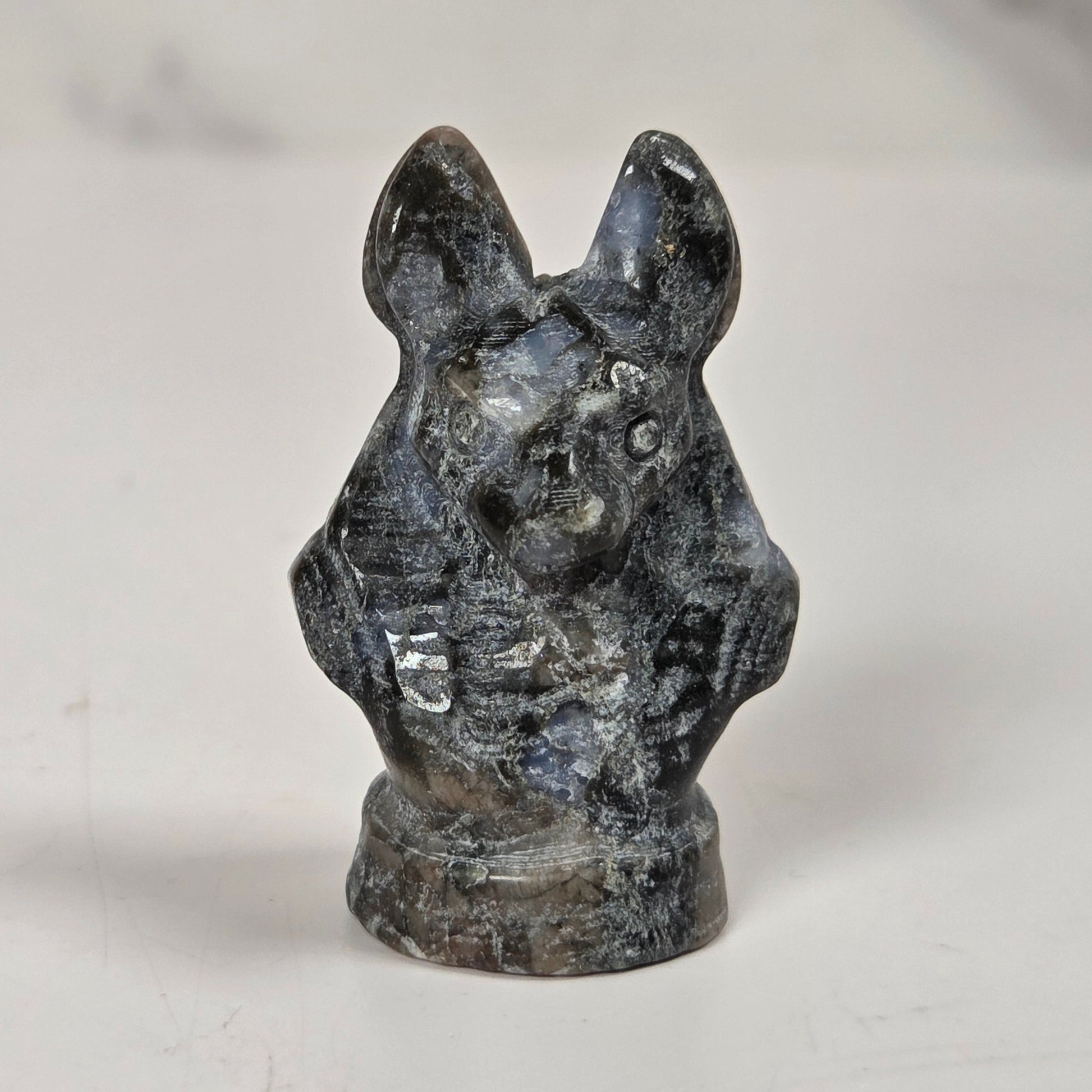 A small carved figurine of a rabbit or hare head and upper body, made from a dark polished stone with gray, black, and blue-gray marbling, standing on a white surface.