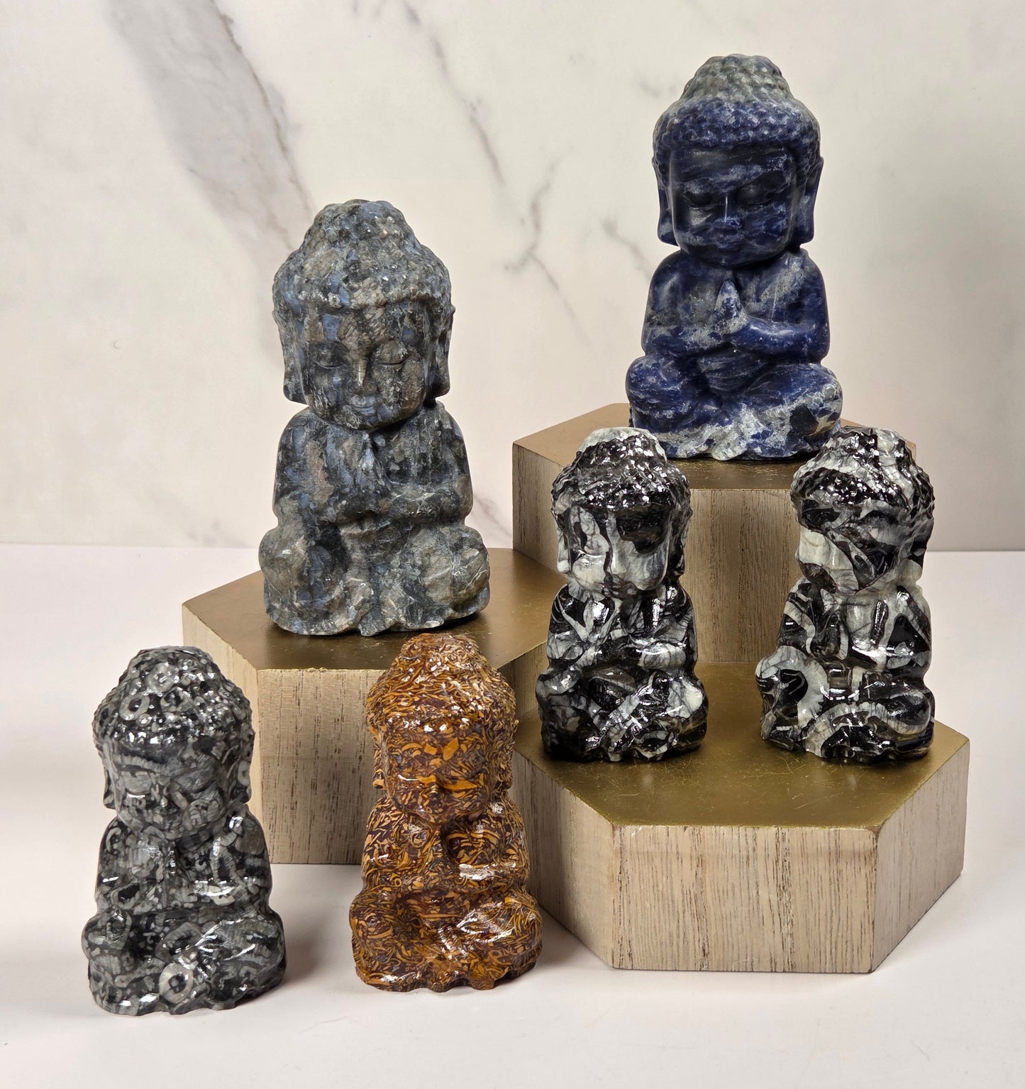 Buddha stone statues in various marbled colors are arranged on wooden display blocks against a light marble-patterned background.