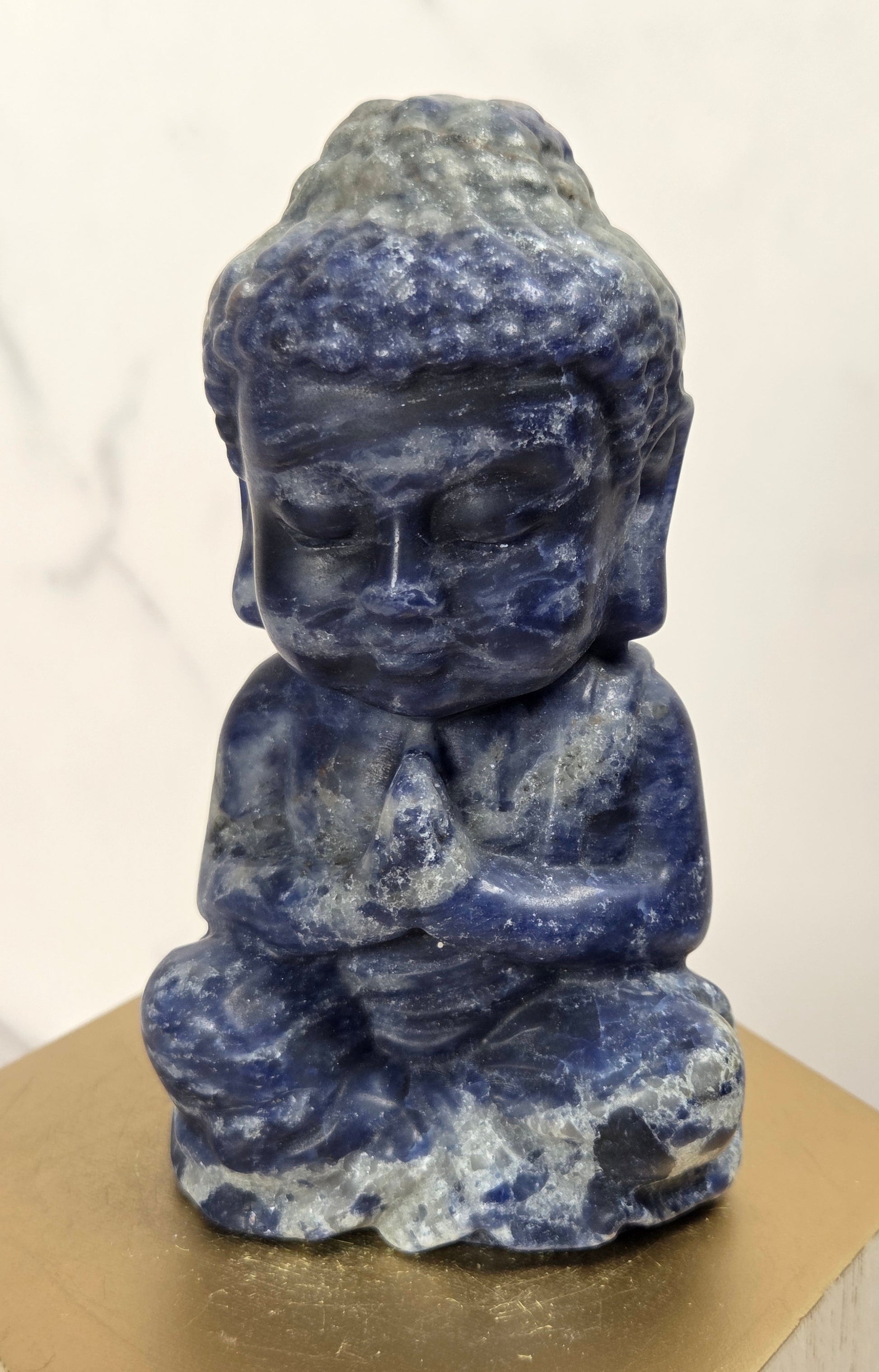 A small meditating Buddha statue carved from blue lapis lazuli, displayed on a gold hexagonal pedestal against a light marble background.