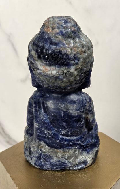 A small carved Buddha statue made of blue and white marbled stone, displayed on a gold hexagonal pedestal against a light marble-patterned background.