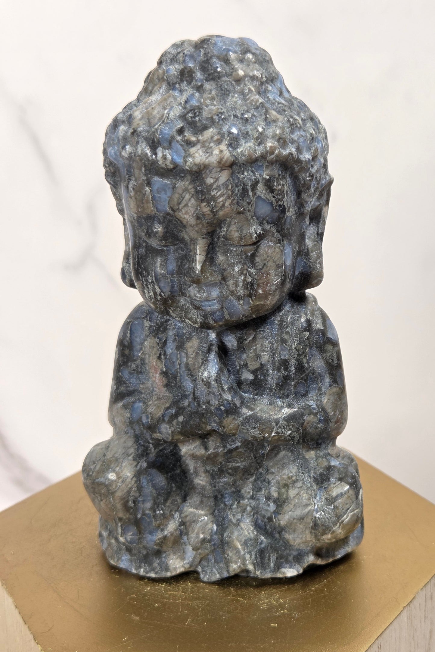 A small carved Buddha statue made of polished labradorite stone, featuring natural blue and gray flashes, displayed on a gold hexagonal pedestal.