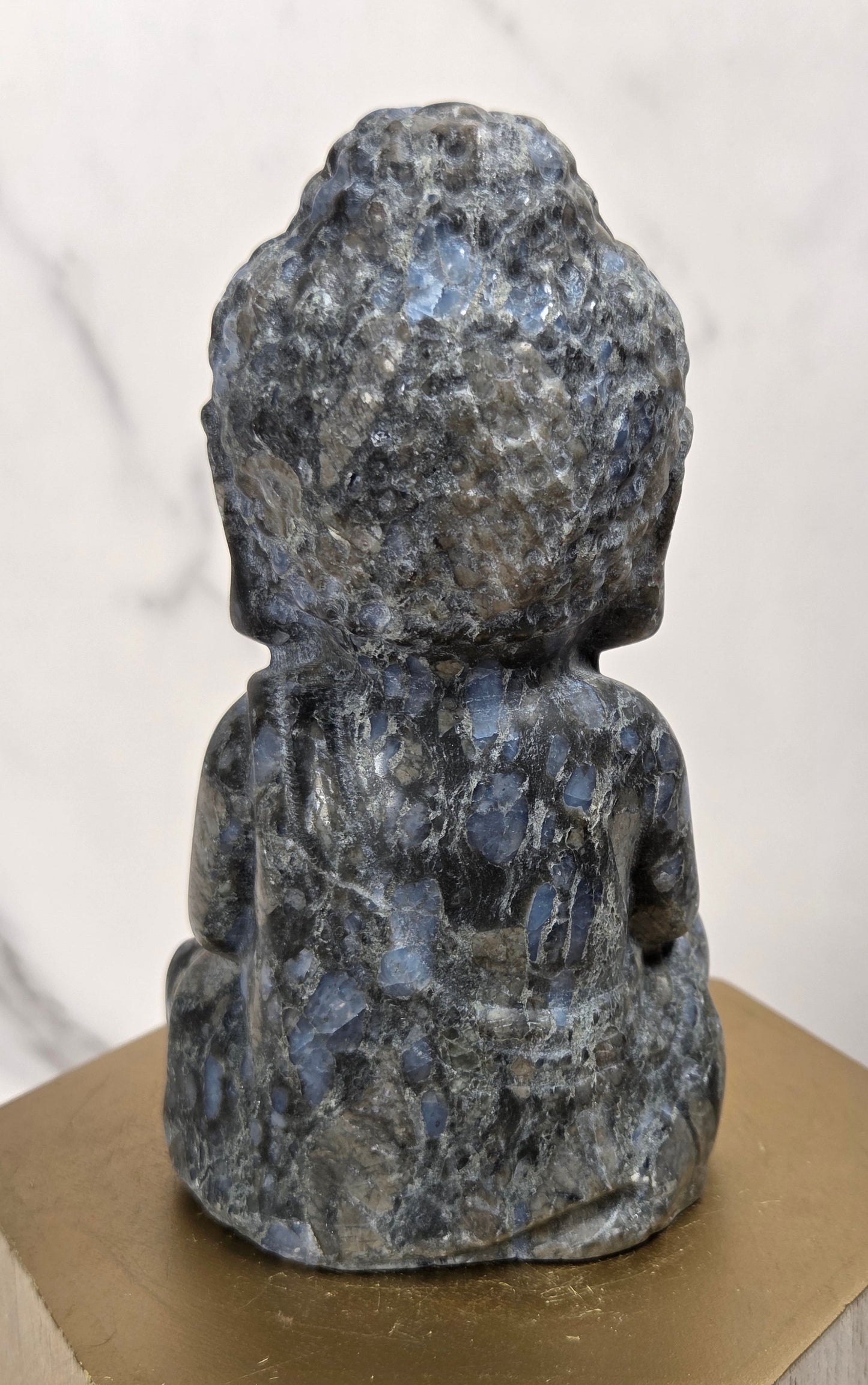 A small carved Buddha statue made of polished blue-gray lapel stone, shown from the back, resting on a hexagonal gold-toned pedestal.