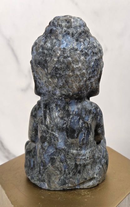 A small carved Buddha statue made of polished blue-gray lapel stone, shown from the back, resting on a hexagonal gold-toned pedestal.