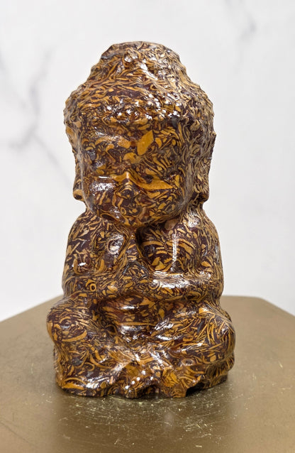 A small Buddha figurine carved from brown and gold patterned wood, sitting on a hexagonal gold-colored pedestal against a white marble background.