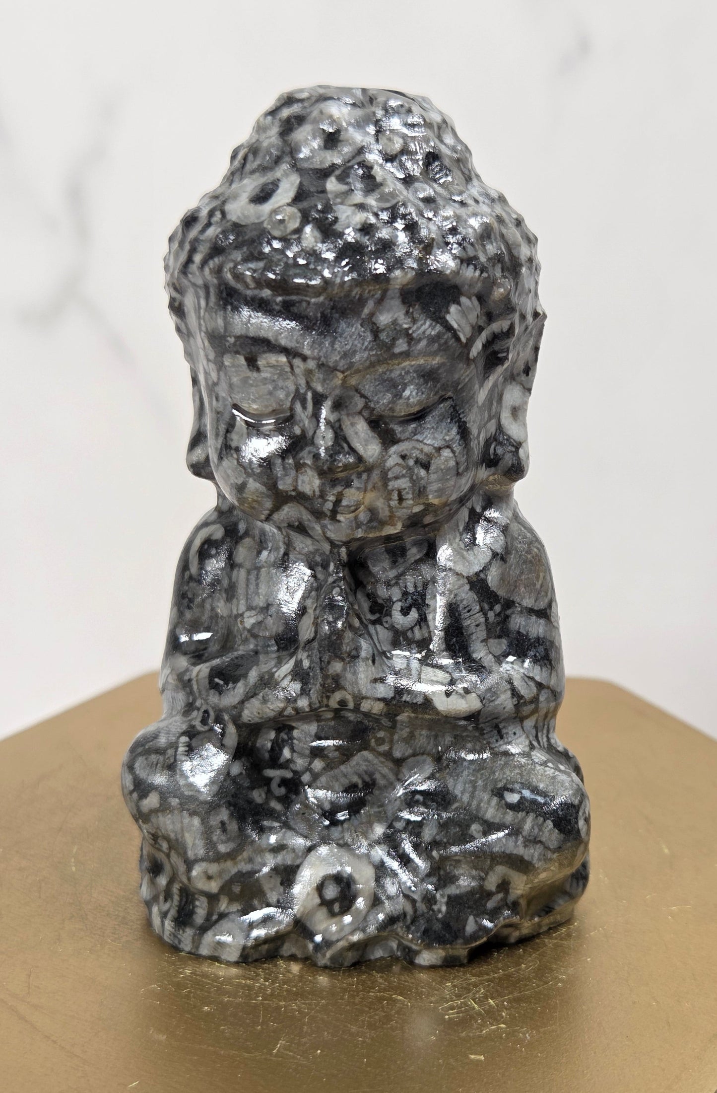 A small black and white marbled figurine of a seated Buddha-like figure, displayed on a gold hexagonal pedestal against a light marble-patterned background.