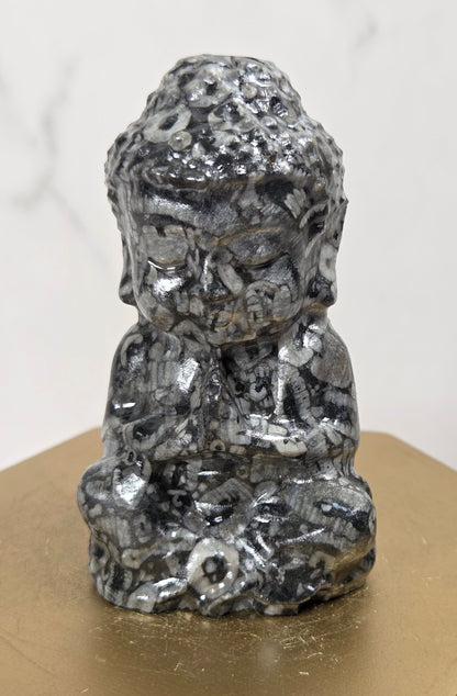 A small black and white marbled figurine of a seated Buddha-like figure, displayed on a gold hexagonal pedestal against a light marble-patterned background.