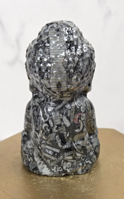 A small sculptural figurine with a textured, metallic silver and black surface, displayed on a gold-colored geometric pedestal against a light marble-patterned background.