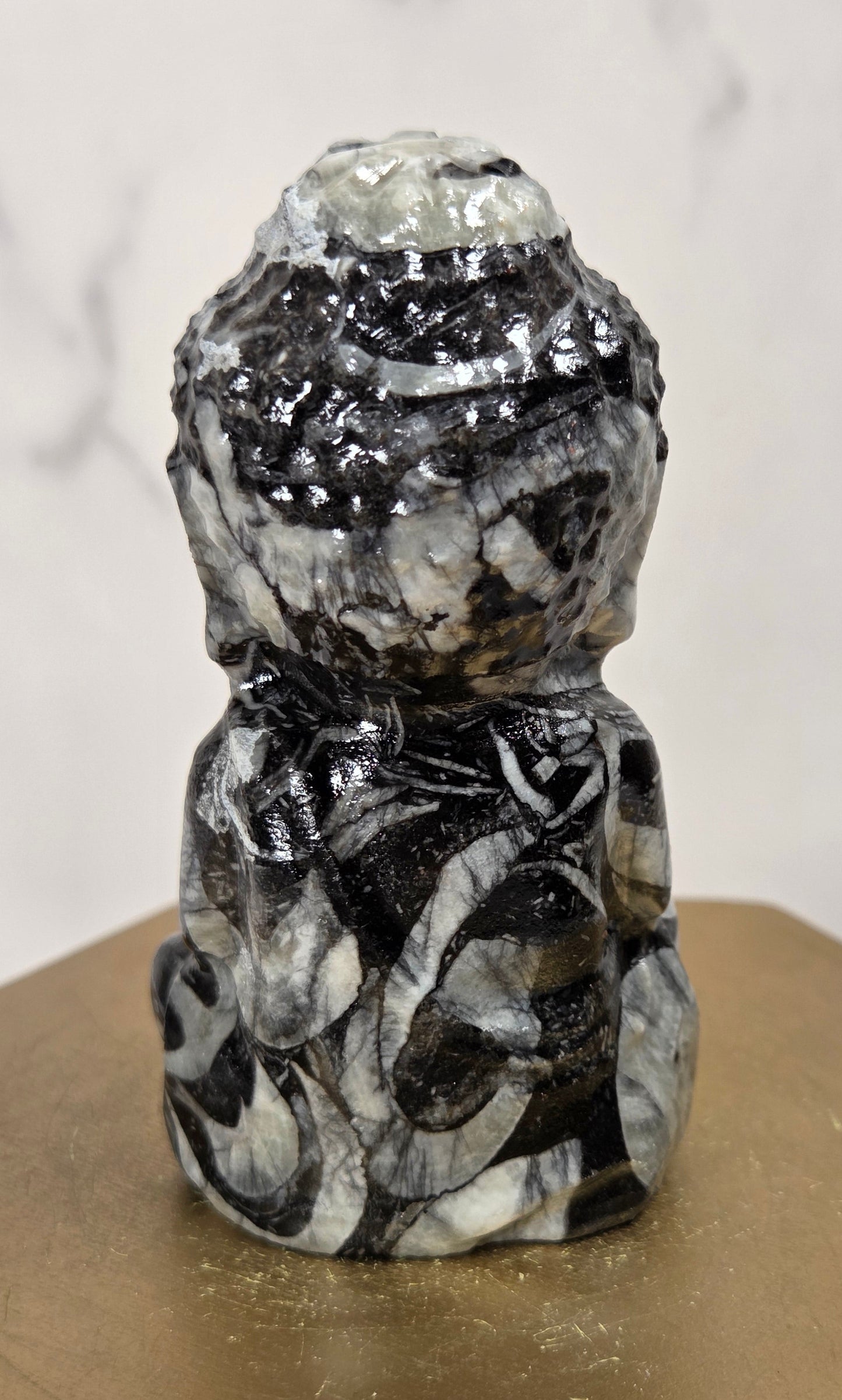 A small carved stone Buddha figurine with a black, white, and gray marbled pattern, displayed on a gold-colored hexagonal block against a light marble background.