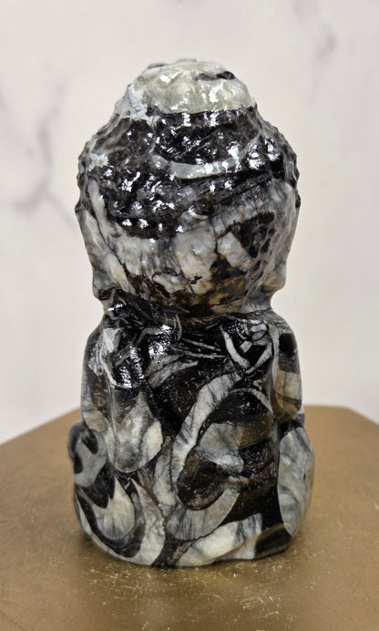 A small carved stone Buddha figurine with a black, white, and gray marbled pattern, displayed on a gold-colored hexagonal block against a light marble background.