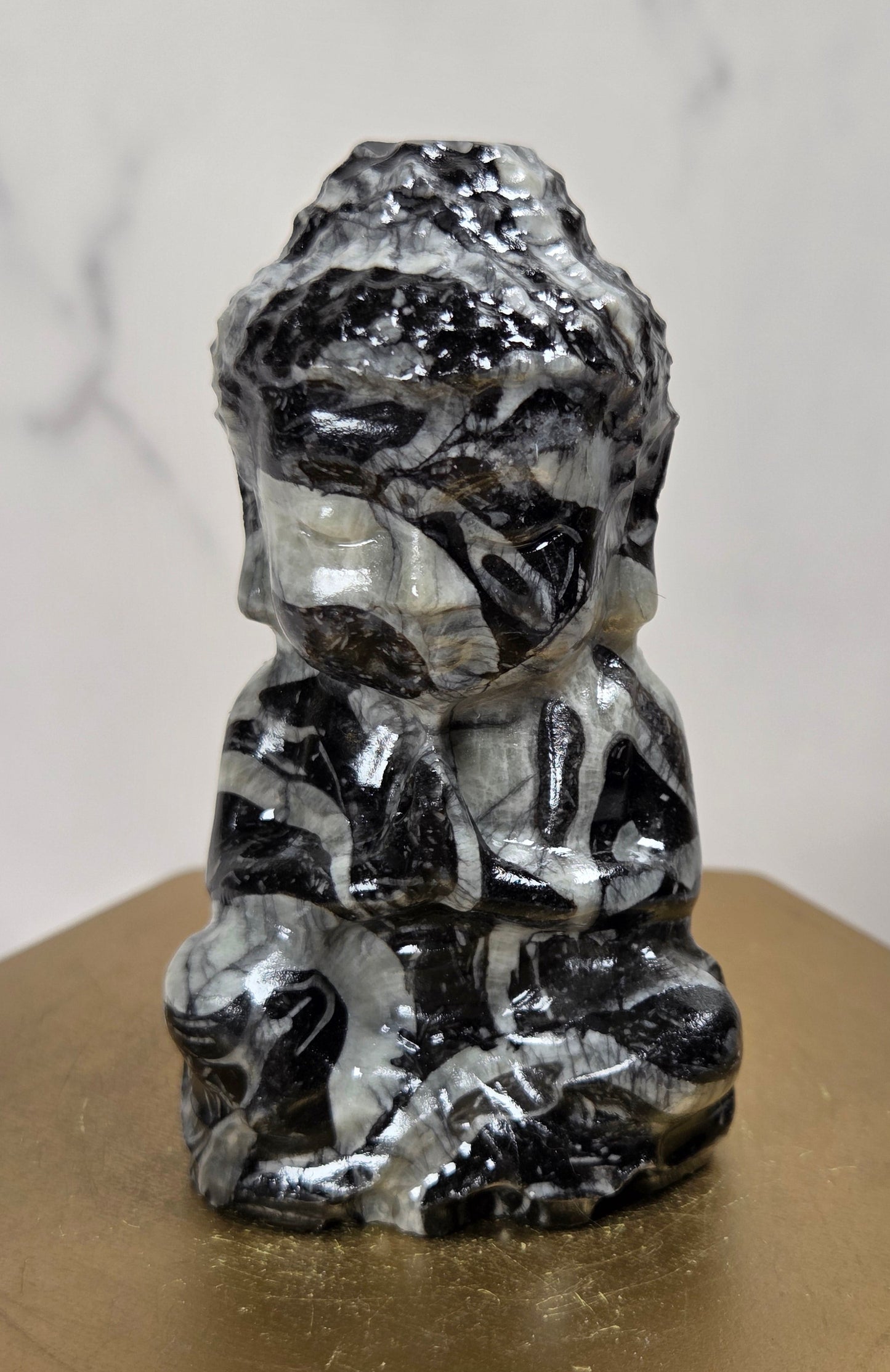 A small carved figurine of a seated Buddha-like figure made from black and white marbled stone, displayed on a gold-colored hexagonal pedestal against a light marble-patterned background.