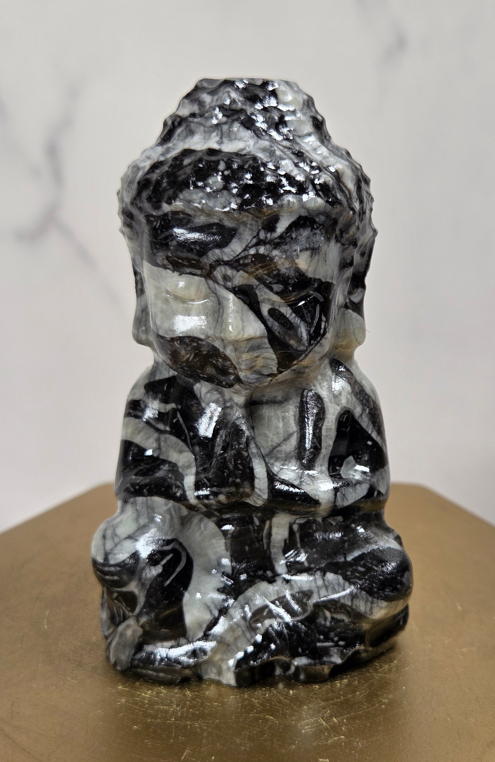 A small carved figurine of a seated Buddha-like figure made from black and white marbled stone, displayed on a gold-colored hexagonal pedestal against a light marble-patterned background.