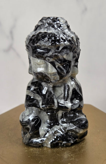 A small carved figurine of a seated Buddha-like figure made from black and white marbled stone, displayed on a gold-colored hexagonal pedestal against a light marble-patterned background.