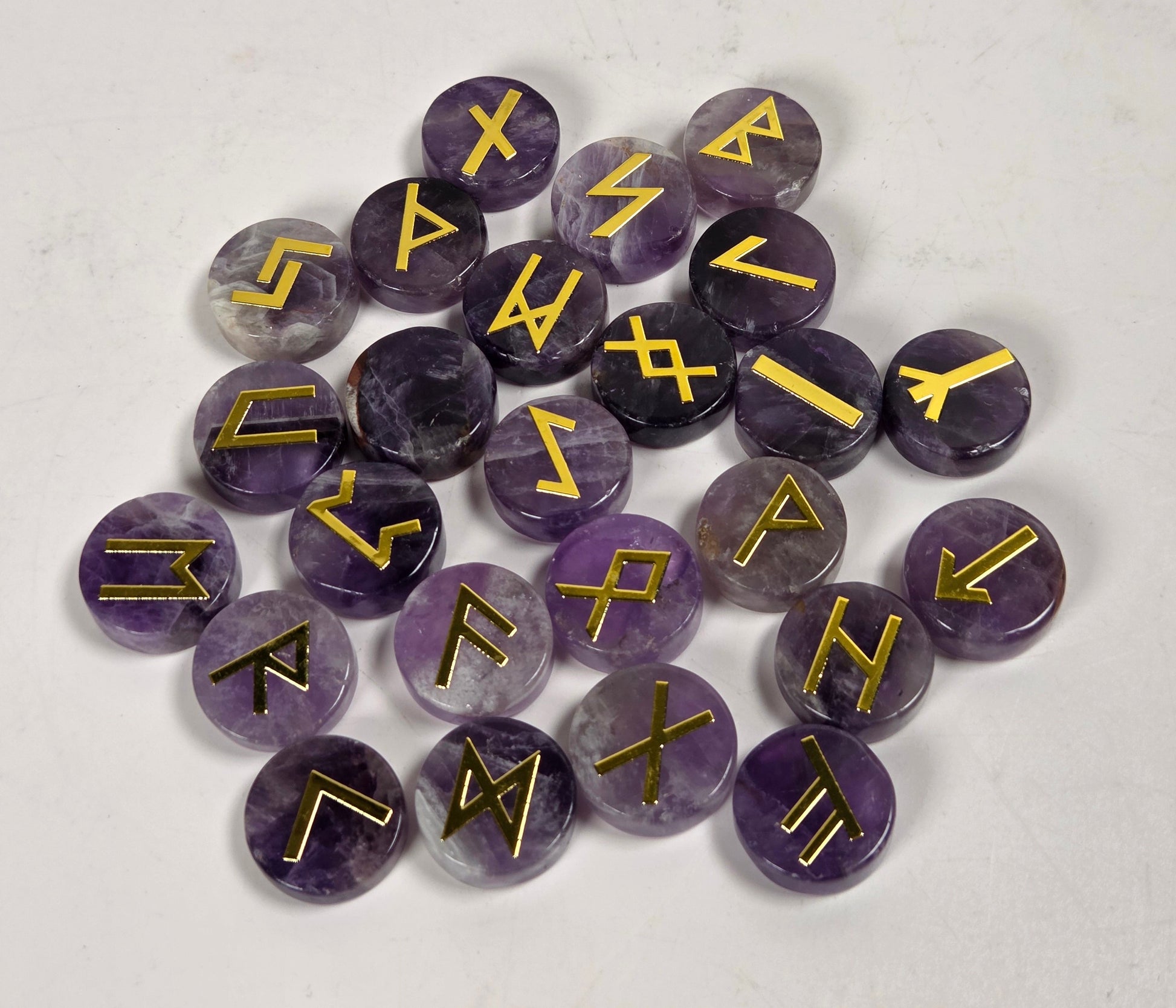 A pile of round purple amethyst cabochons, each engraved with a different gold rune symbol, arranged on a light gray textured surface.