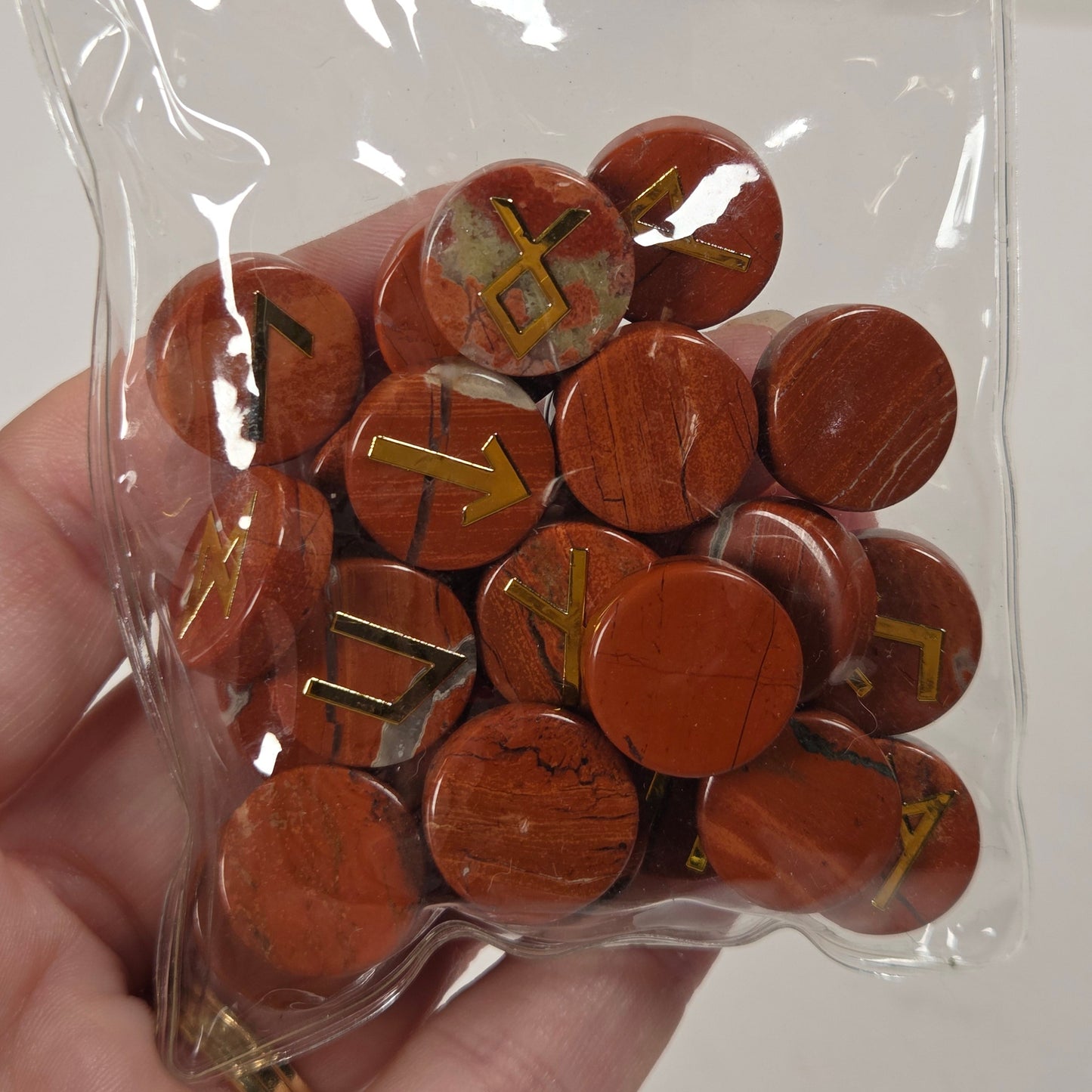 A hand holding a clear plastic bag filled with round reddish-brown wooden tokens or discs, some marked with yellow symbols, against a plain light background.
