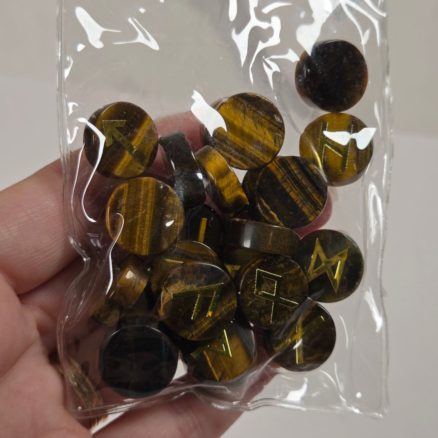 A hand holding a clear plastic bag filled with round, polished dark brown and black gemstone beads featuring engraved gold-colored symbols, set against a light gray background.