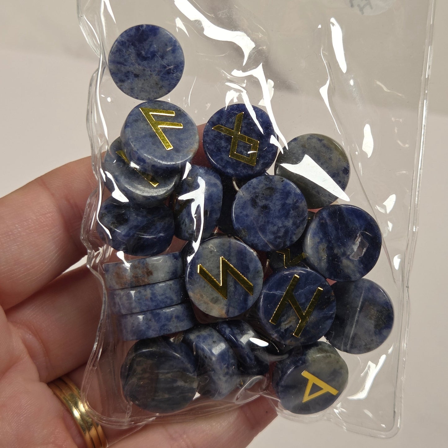 A hand holding a clear plastic bag filled with round blue stone pendants featuring engraved gold runes, against a light neutral background.