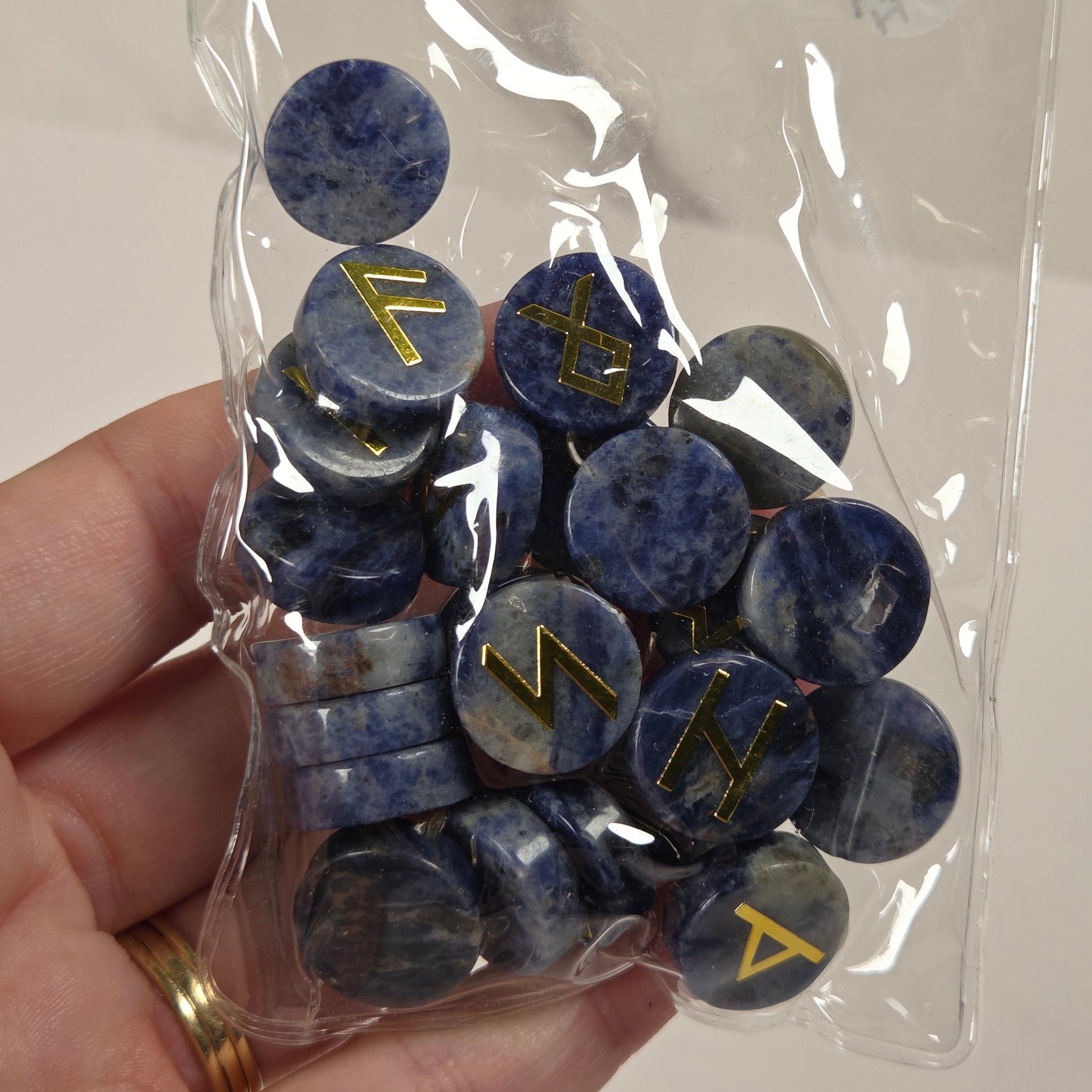 A hand holding a clear plastic bag filled with round blue stone pendants featuring engraved gold runes, against a light neutral background.