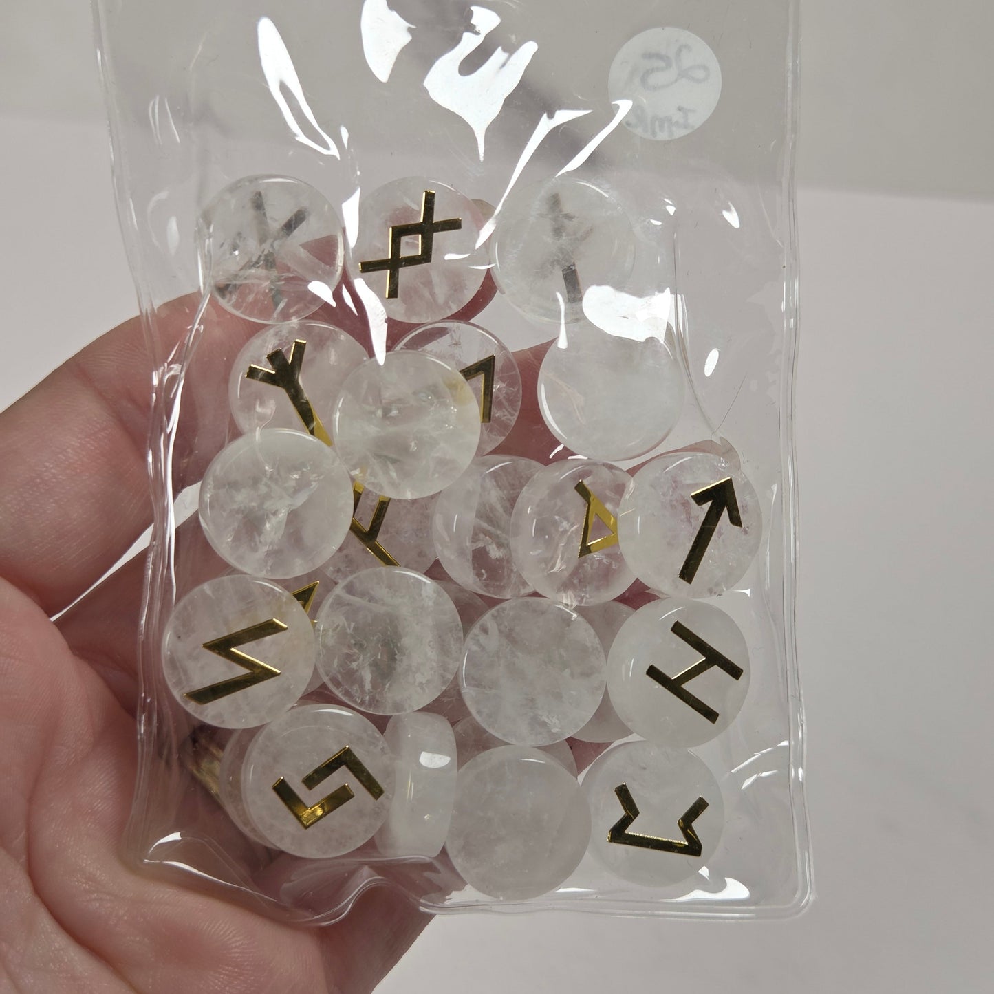 A hand holding a clear plastic zip-top bag filled with round, translucent white cabochon stones, each stamped with a black directional arrow or symbol. A small white circular sticker with the brand name 'The Selenite Shop' is visible on the bag.