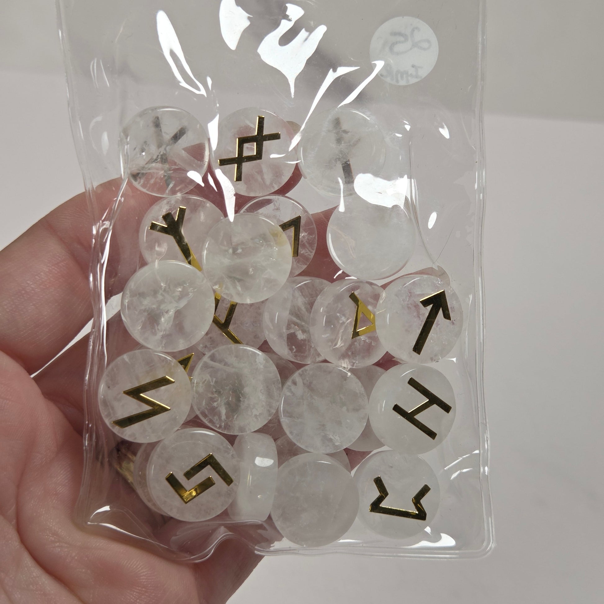 A hand holding a clear plastic zip-top bag filled with round, translucent white cabochon stones, each stamped with a black directional arrow or symbol. A small white circular sticker with the brand name 'The Selenite Shop' is visible on the bag.