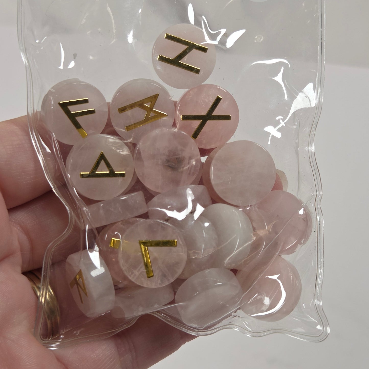 A hand holding a clear plastic bag filled with round pale pink crystal beads, each engraved with a small gold rune symbol, against a plain light background.