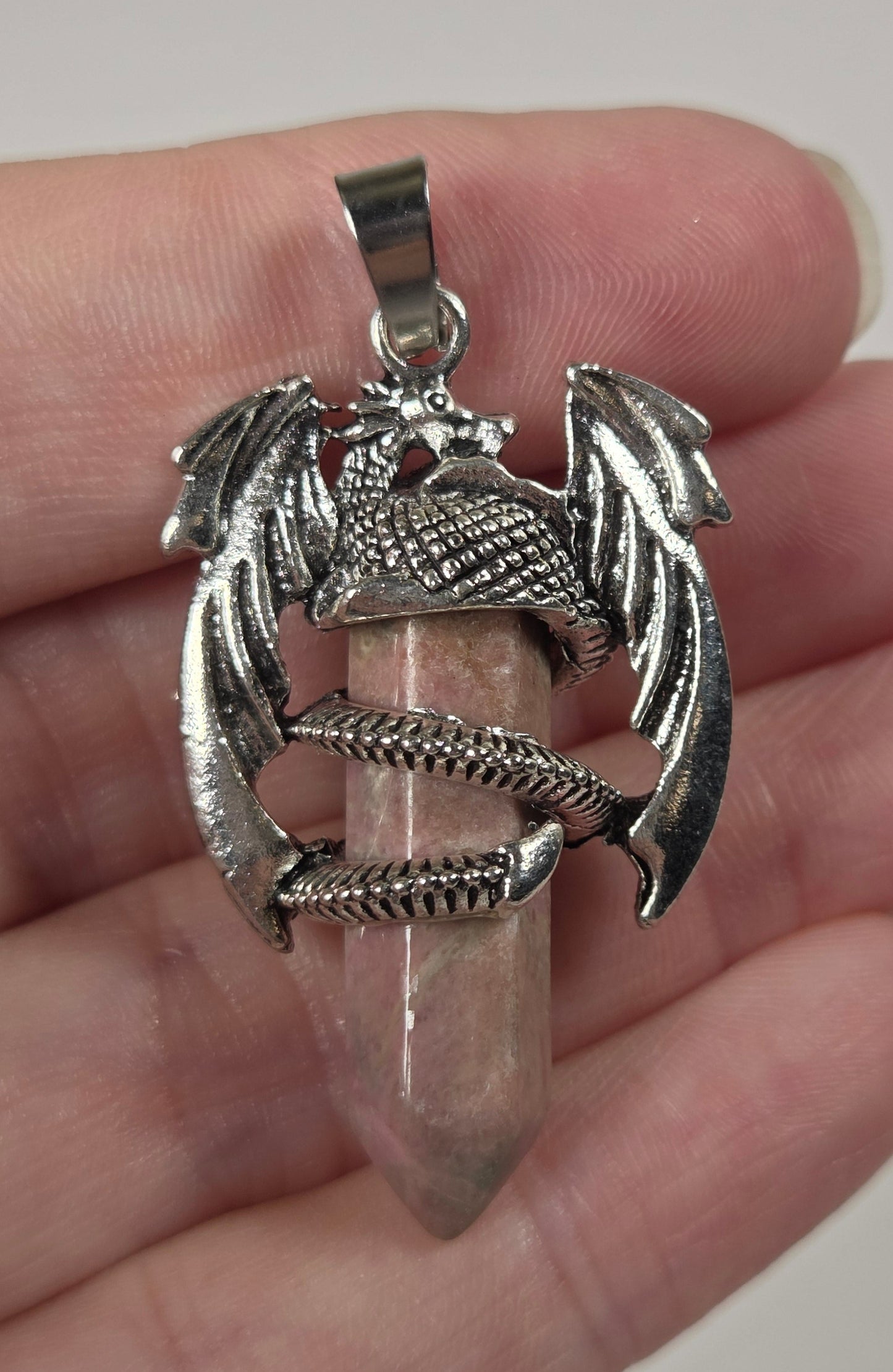 A silver-toned pendant featuring a detailed dragon clutching a pinkish crystal point, held in a person's hand against a plain light background.