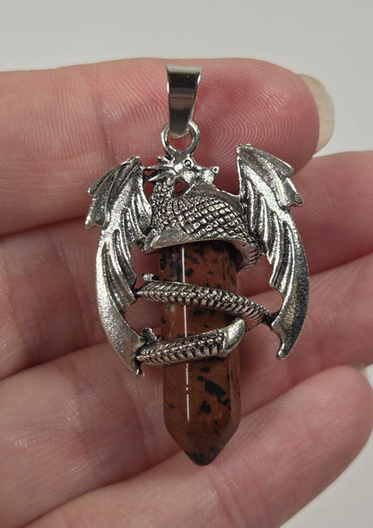 A hand holding a silver-toned dragon pendant wrapped around a brown speckled stone point, with a gold ring on the left hand.