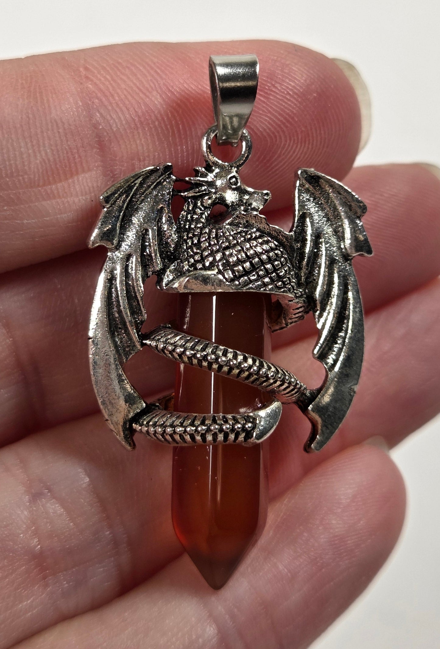 A hand holding a silver dragon pendant wrapped around a pointed amber-red gemstone, against a plain light background.