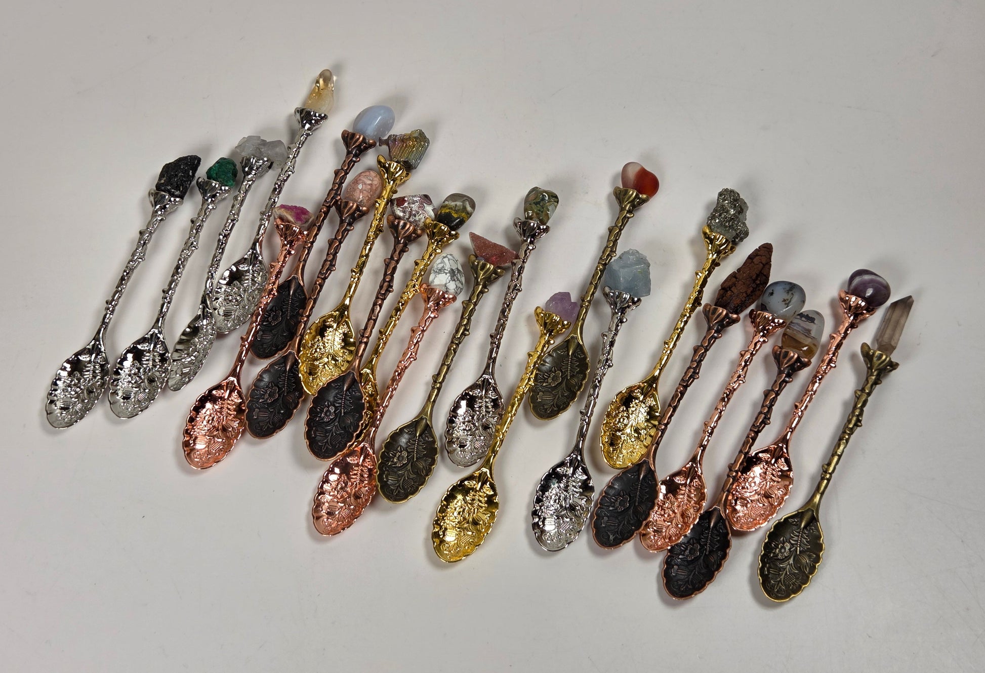 A group of decorative miniature spoons arranged in a fan shape on a plain light background. The spoons have textured metallic handles and bowls in various finishes including silver, gold, rose gold, bronze, and dark gunmetal, with some featuring colorful gemstone accents.