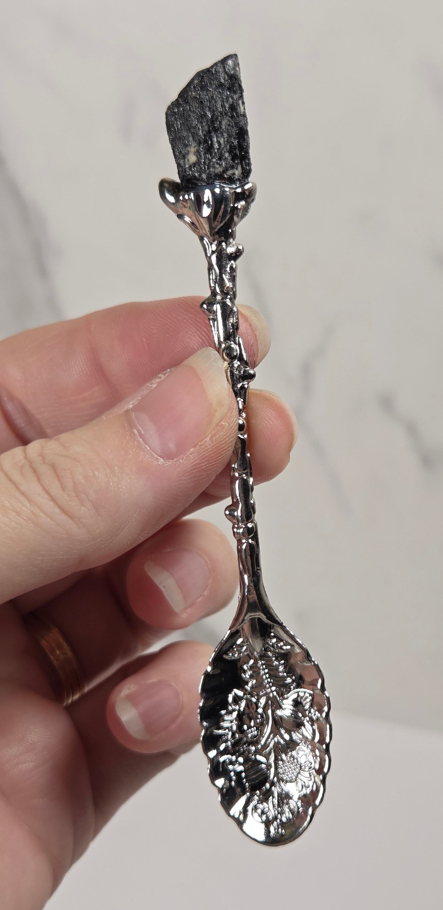 A hand holding a decorative silver spoon with a textured, hammered finish and a dark, polished stone-like top piece, set against a light gray marble-patterned background.