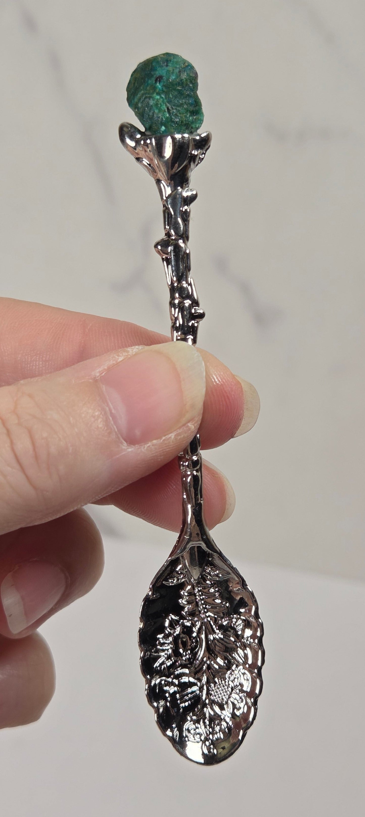 A hand holding a decorative silver spoon with a textured leaf-shaped bowl and a small green gemstone at the top, set against a light gray marble background.