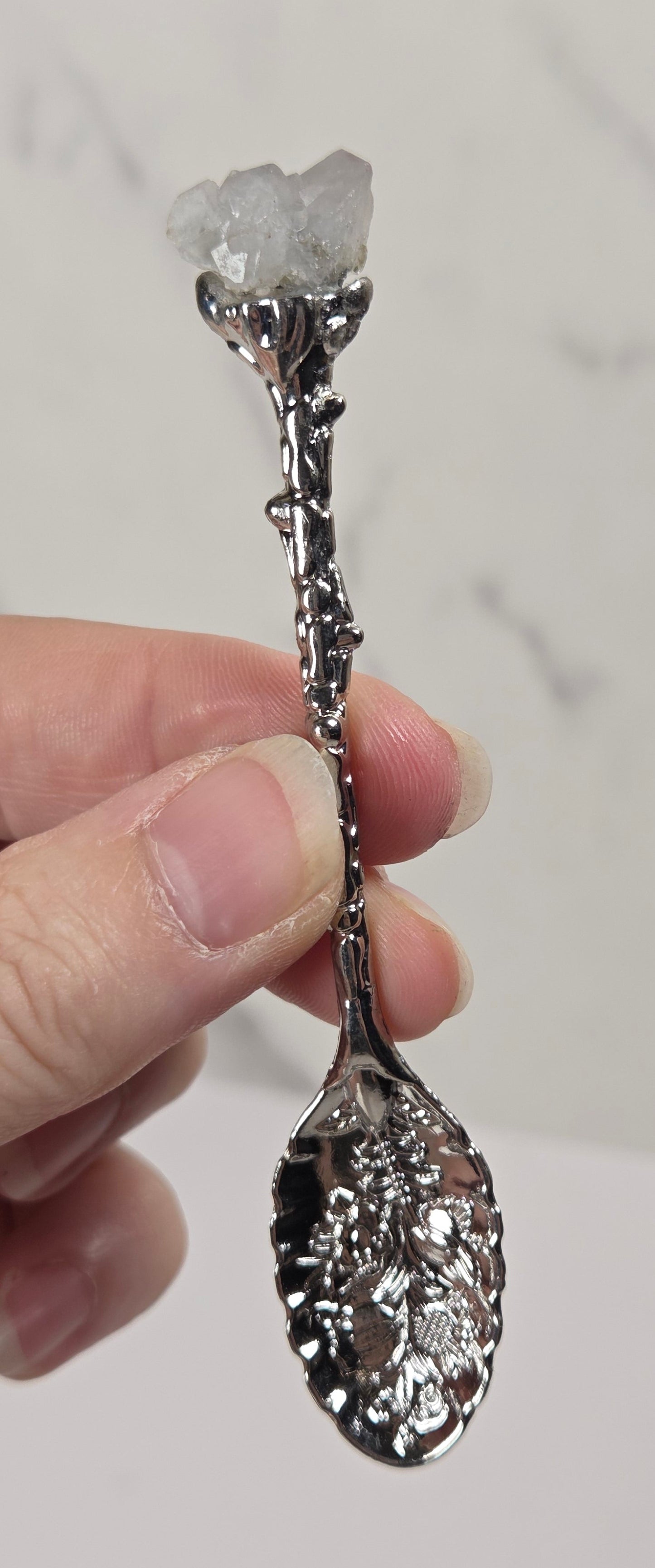 A hand holding a decorative silver spoon with a clear crystal cluster at the bowl, set against a light marble-patterned background.