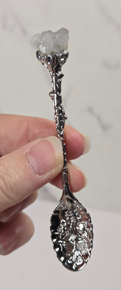 A hand holding a decorative silver spoon with a clear crystal cluster at the bowl, set against a light marble-patterned background.