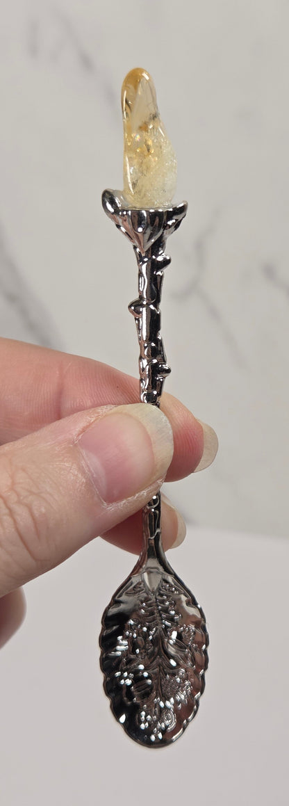 A hand holding a decorative spoon with a dark metallic leaf-shaped bowl and stem, topped with a small amber crystal or glass piece, against a light marble-patterned background.