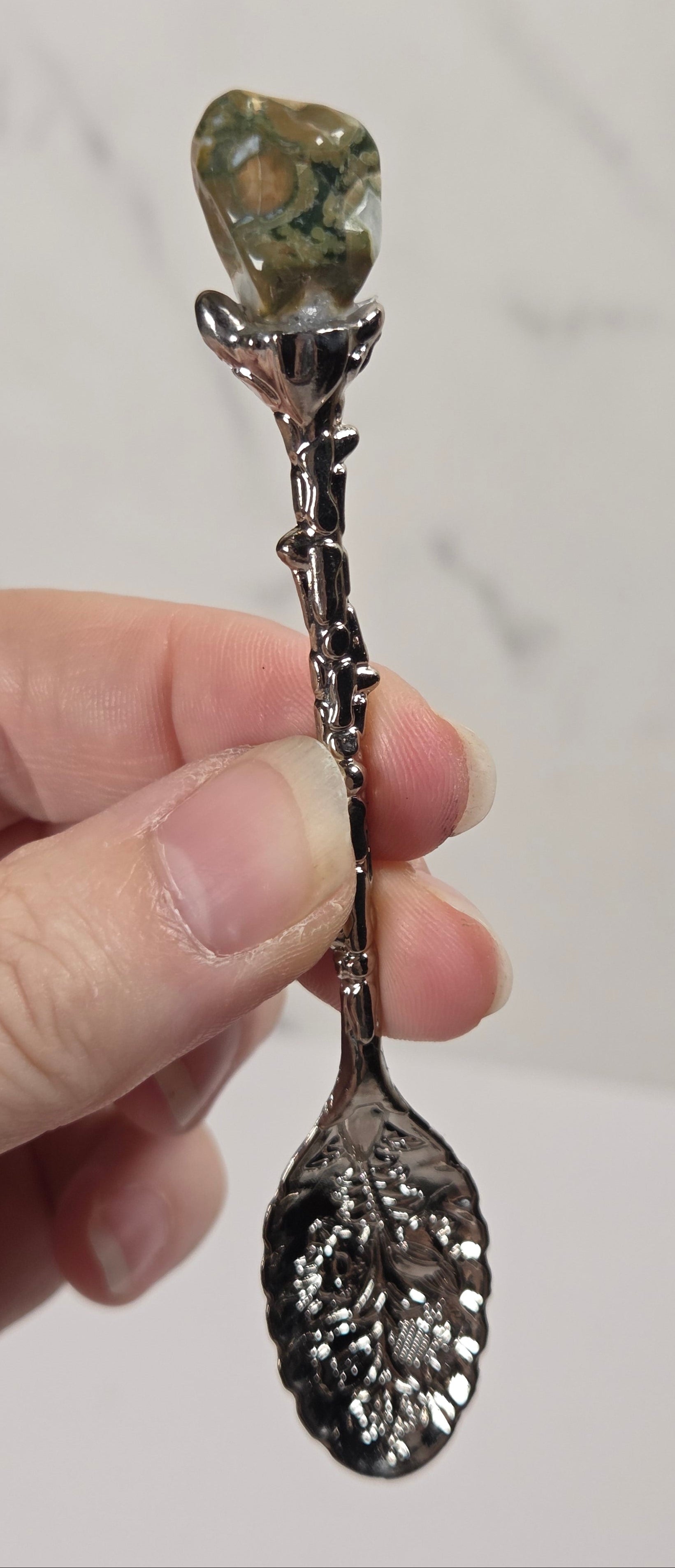 A hand holding a decorative spoon with an ornate silver-toned stem and a polished green stone set at the top, against a light marble-patterned background.
