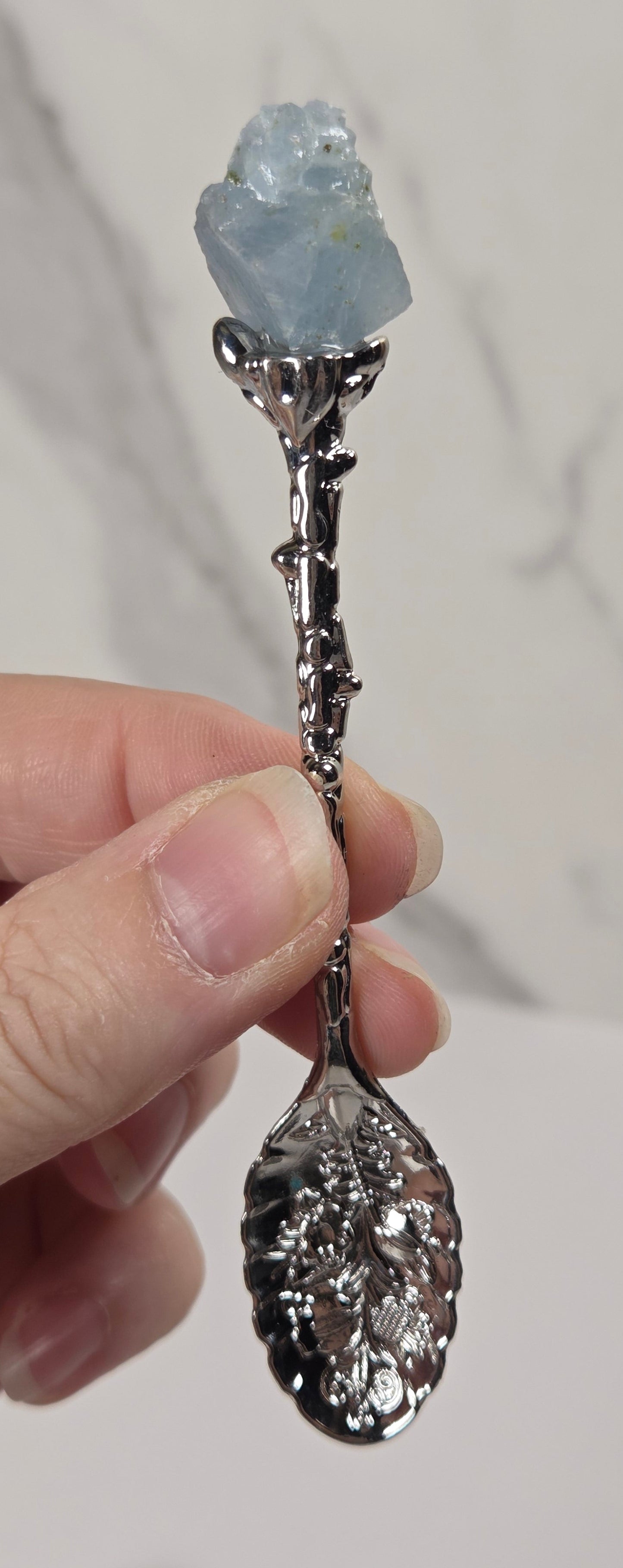 A hand holding a decorative spoon with a polished silver leaf-shaped bowl and stem, topped with a raw light blue gemstone, against a light gray marble background.