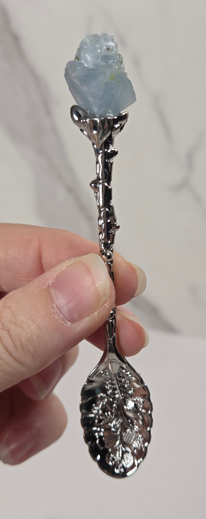 A hand holding a decorative spoon with a polished silver leaf-shaped bowl and stem, topped with a raw light blue gemstone, against a light gray marble background.