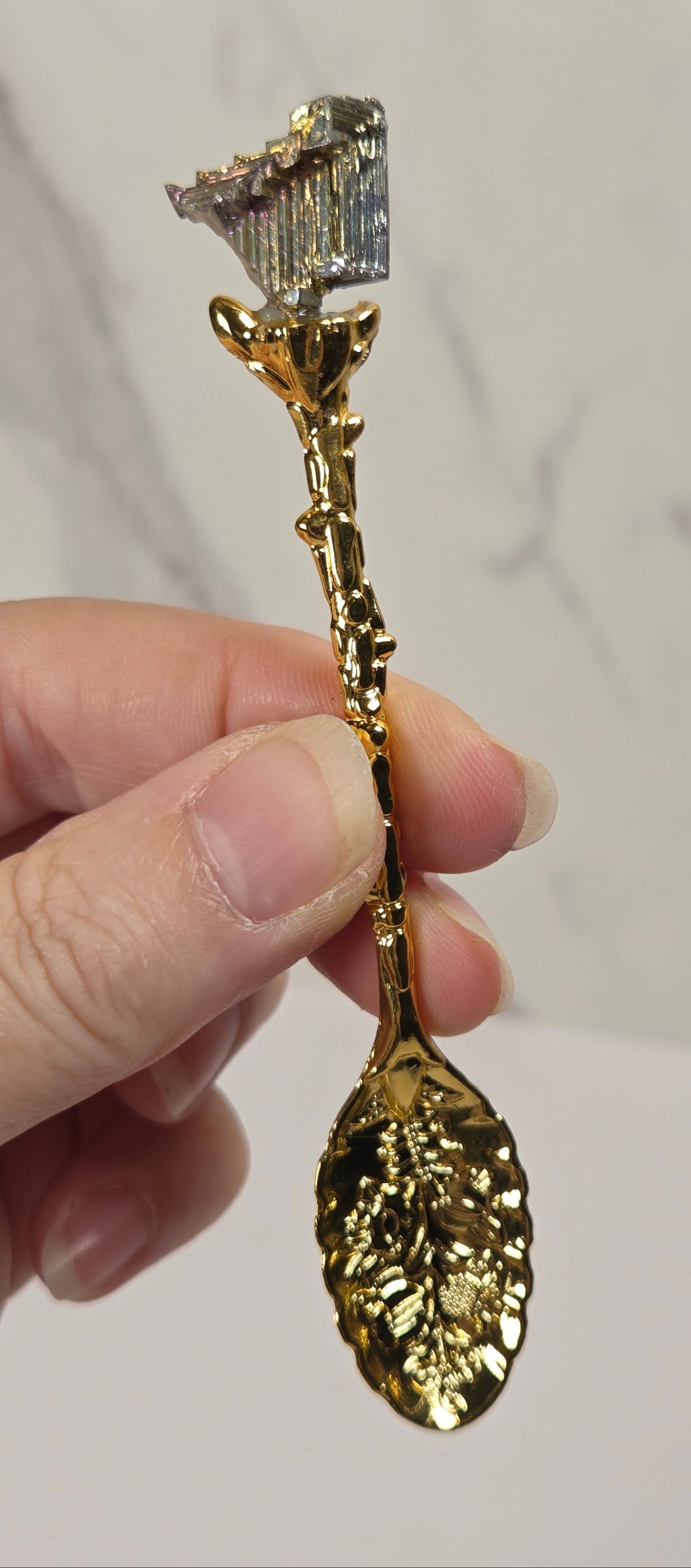 A hand holding a decorative gold spoon with a leaf-shaped bowl and a small crystal cluster at the top, set against a light marble-patterned background.