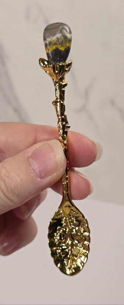 A hand holding a decorative gold spoon with a leaf-shaped bowl and a stem that resembles a flower bud with a clear, patterned top, set against a light marble background.