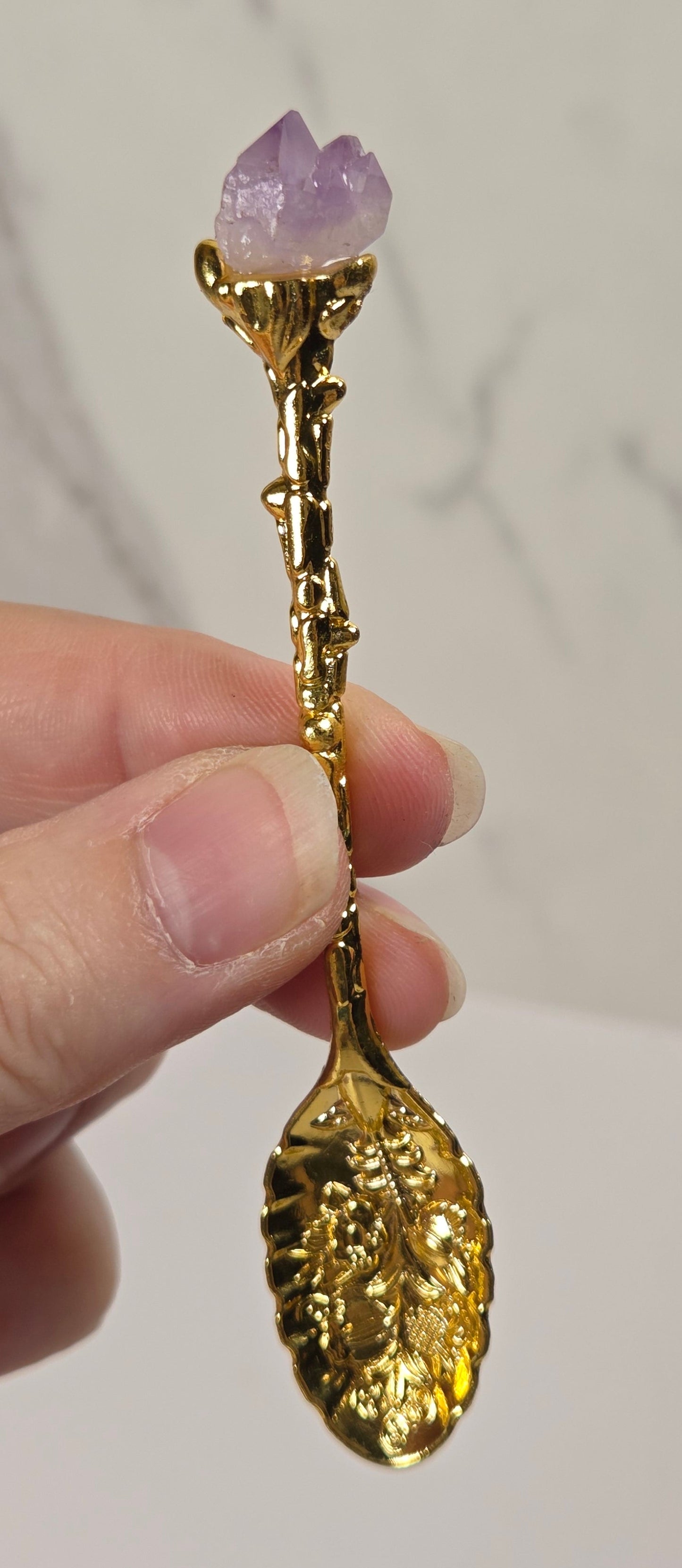 A hand holding a decorative gold spoon with a textured stem and a purple crystal gemstone at the top, set against a light gray marble-patterned background.