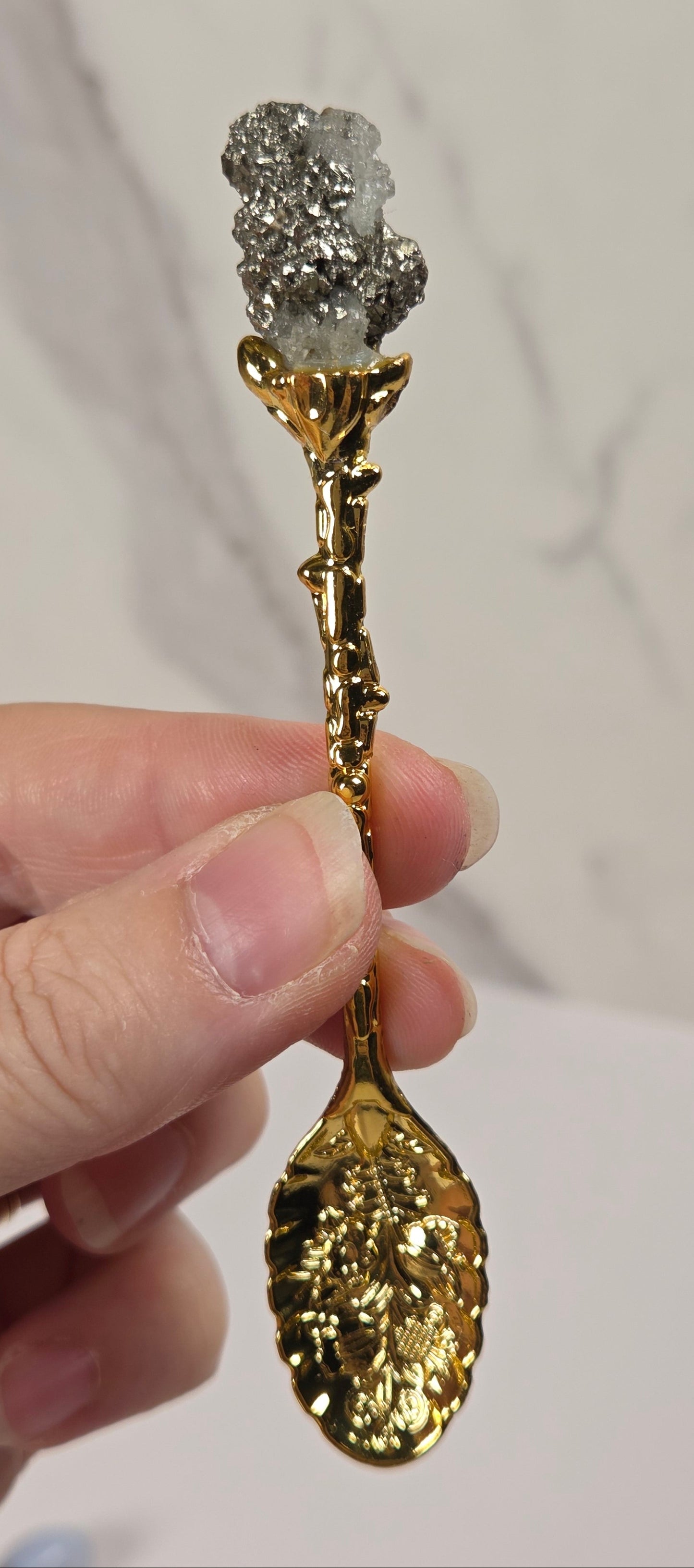 A hand holding a decorative gold spoon with a textured crystal cluster at the bowl, set against a light marble-patterned background.