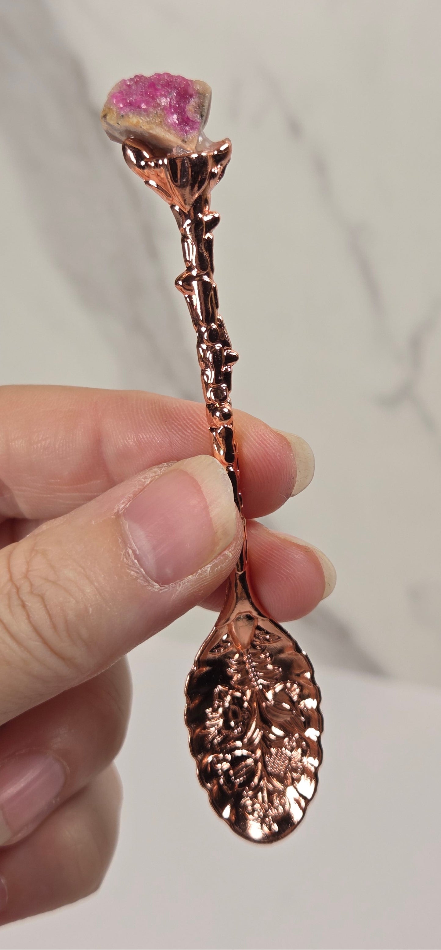 A hand holding a decorative rose-shaped spoon or utensil with a metallic rose-gold finish and a pink gemstone flower head, against a light marble-patterned background.