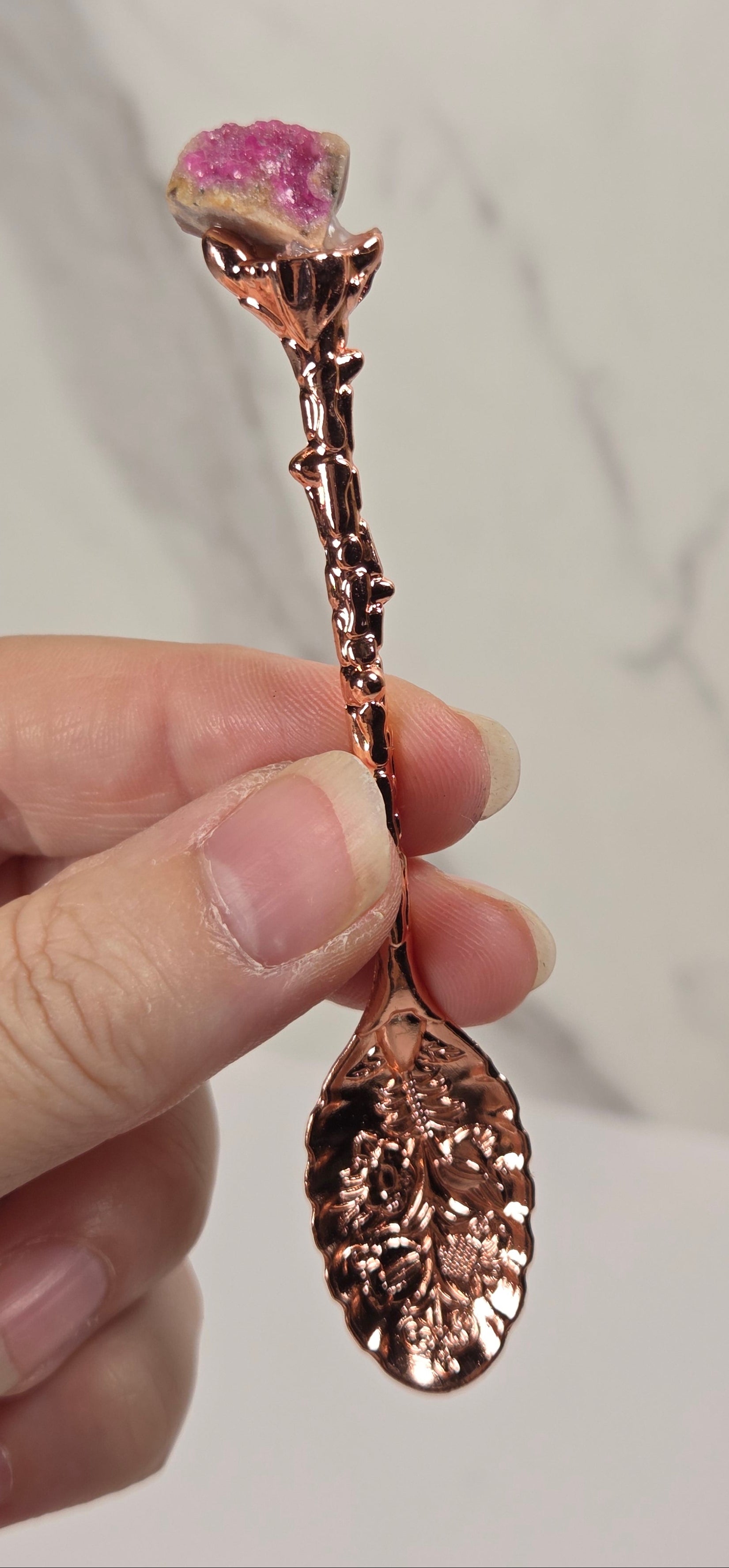 A hand holding a decorative rose-shaped spoon or utensil with a metallic rose-gold finish and a pink gemstone flower head, against a light marble-patterned background.