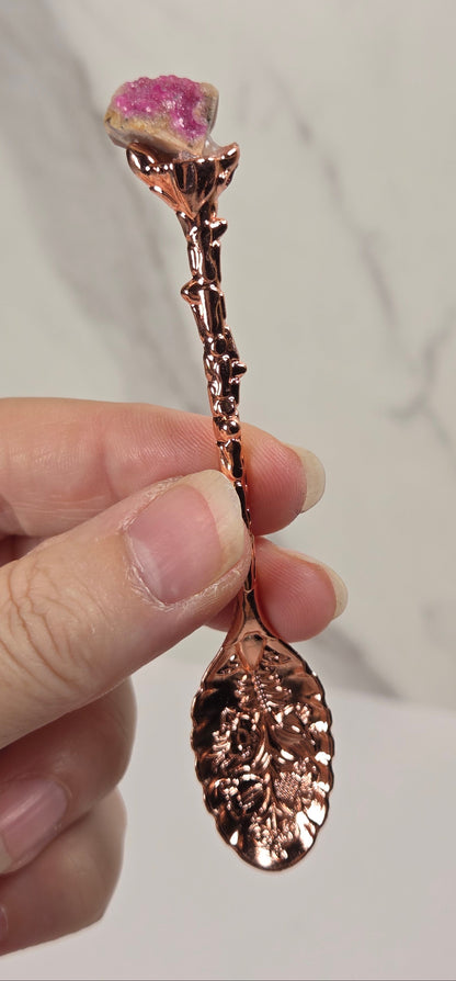A hand holding a decorative rose-shaped spoon or utensil with a metallic rose-gold finish and a pink gemstone flower head, against a light marble-patterned background.