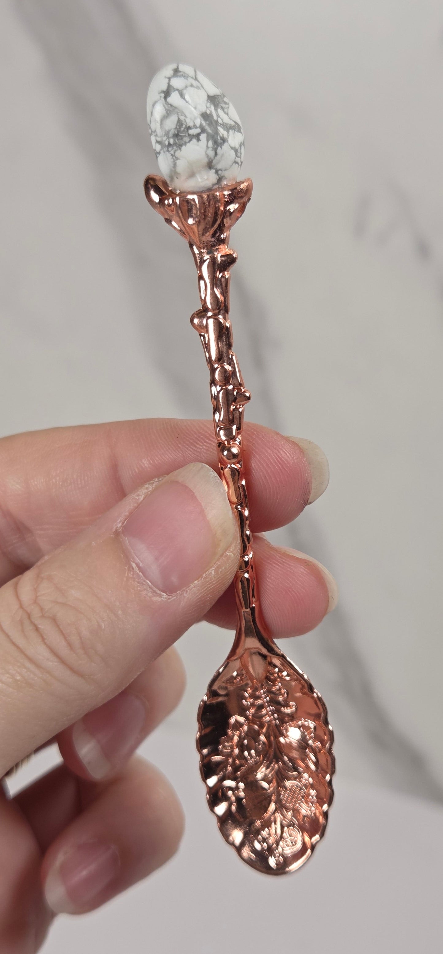 A hand holding a decorative spoon with a rose gold metallic leaf-shaped handle and a white marbled gemstone on the tip, set against a light gray marble background.