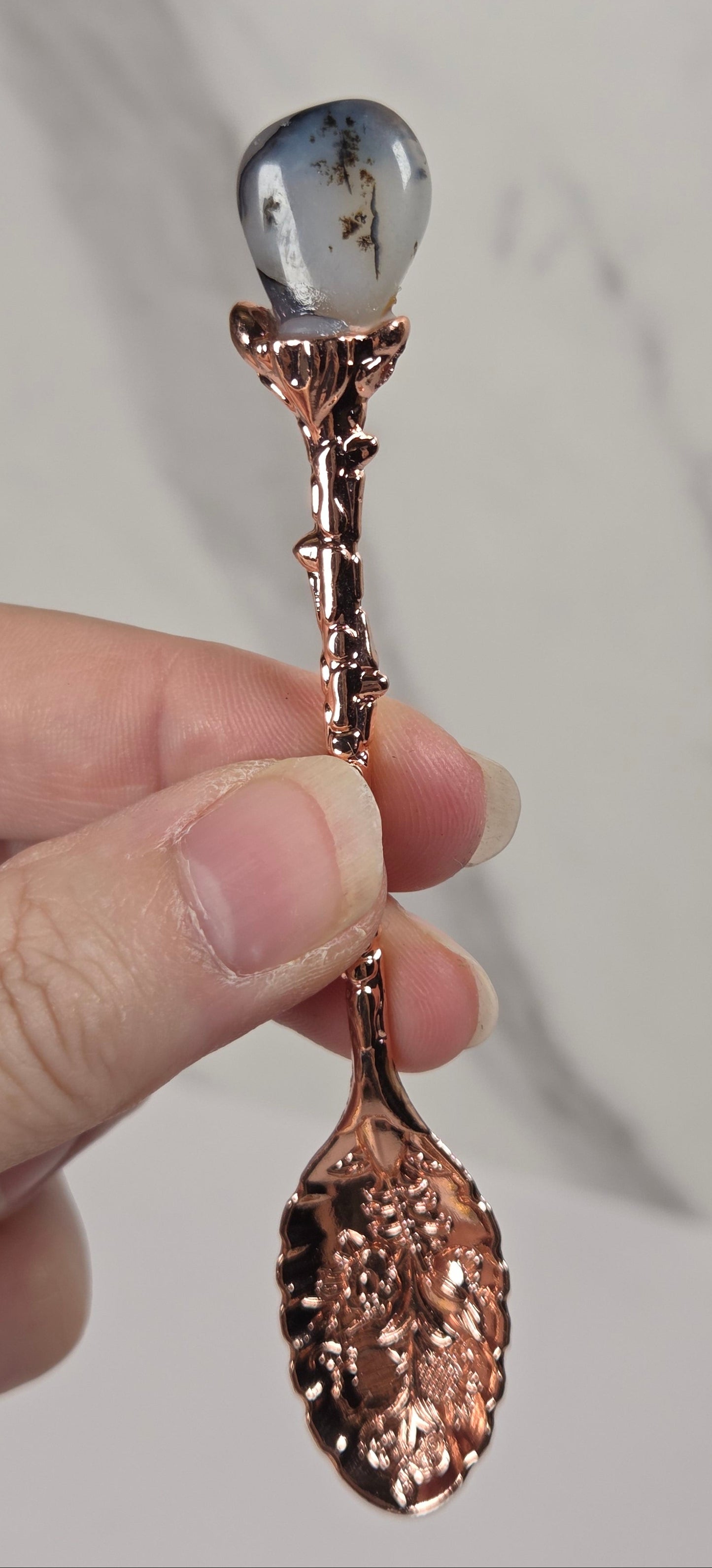 A hand holding a decorative copper spoon with a teardrop-shaped clear stone inlay on the bowl, set against a light gray marble background.