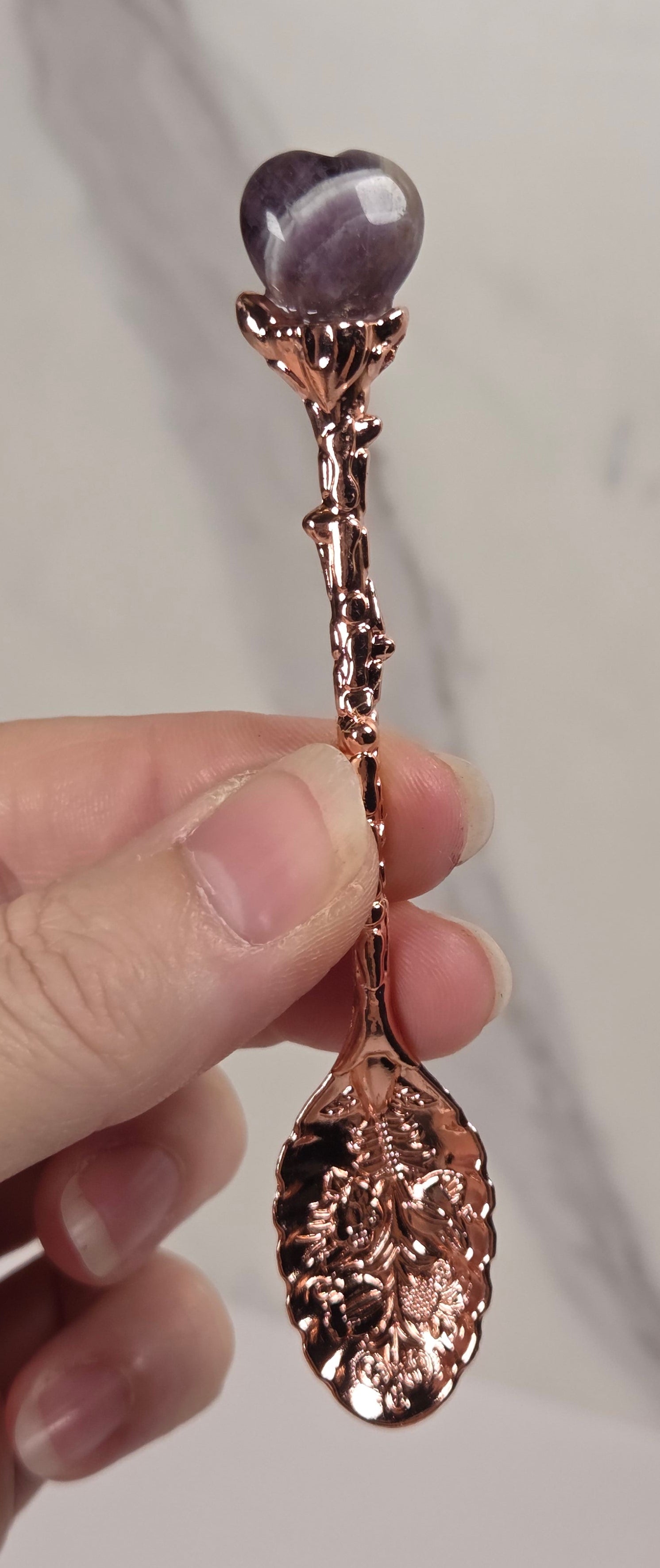 A hand holding a decorative rose gold spoon with a heart-shaped purple gemstone at the top, set against a light marble background.